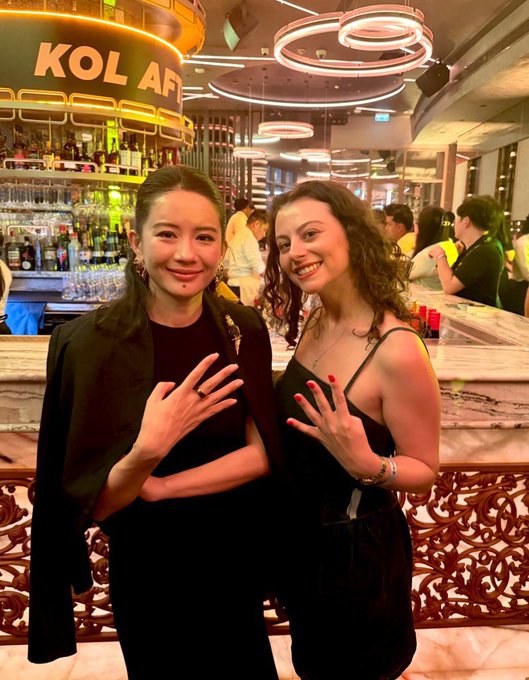 The image features two women standing next to each other, both wearing black dresses. They are posing for a picture together at a bar or restaurant with neon lights illuminating the scene. The woman on the left has her nails painted red, while the other woman also has her nails done in red nail polish. The background of the photo shows various bottles and wine glasses, suggesting that they might be enjoying drinks at the establishment.