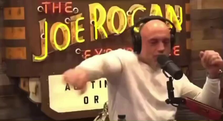 The image features a man wearing headphones, dancing or performing at a microphone studio. He appears to be the host of a radio show called "The RoCAAN", which is known for its humorous content and satirical takes on various topics. The background of the image includes a neon sign that reads "THE ROCAAN". 

Additionally, there are two people in the scene – one man wearing headphones near the microphone and another person standing next to it with their hand on their hip. They might be other radio show hosts or guests involved in this performance or event.