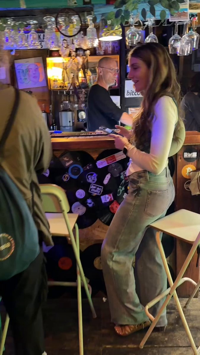 The image shows a woman sitting at a bar, holding a drink and laughing. She has a backpack next to her chair. The bar also features a counter with various drinks displayed behind it, including wine glasses and beer taps. There are several people in the background, some of whom appear to be engaged in conversation or enjoying their time at the bar.