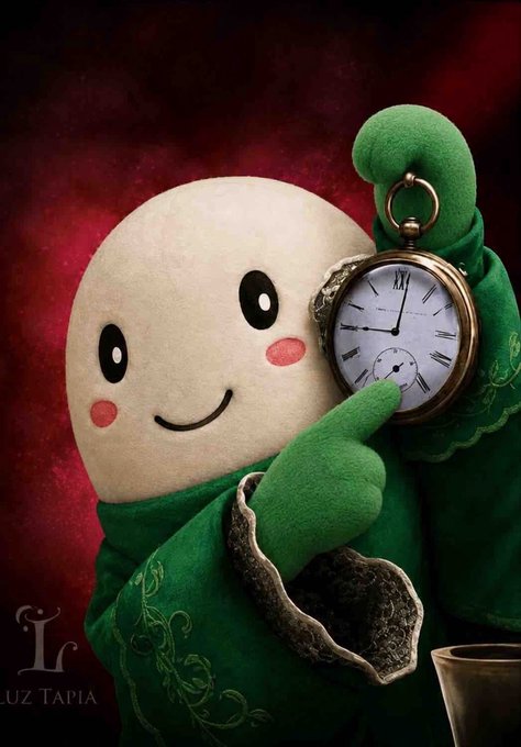 A round, cream-colored plush character with a cheerful smile and pink cheeks, wearing an ornate green embroidered robe, proudly holds up a vintage pocket watch displaying approximately 2:28. The image is watermarked "LUZ TAPIA" with an "L" logo in the bottom corner against a dark red background.