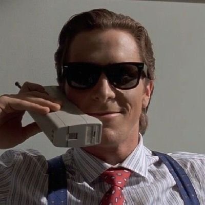 A man with slicked-back brown hair wearing dark sunglasses, a striped shirt, red patterned tie, and suspenders, holding a large retro mobile phone to his ear.
