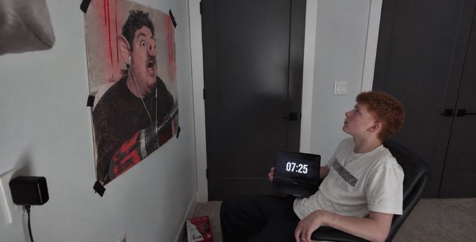 A young man with reddish hair sitting in a chair in a dimly lit room, looking at a poster of a surprised man taped to the wall while holding a tablet showing the time '07:25'.