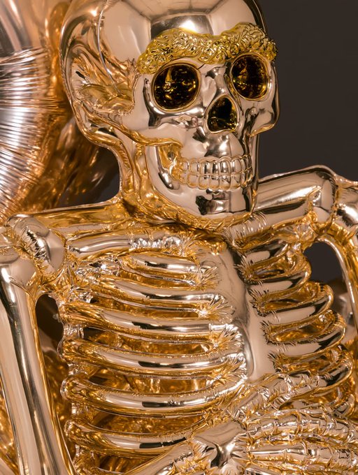 A highly reflective chrome and gold metallic sculpture of a skull and skeletal torso, featuring amber-tinted eye sockets and intricate rib details.