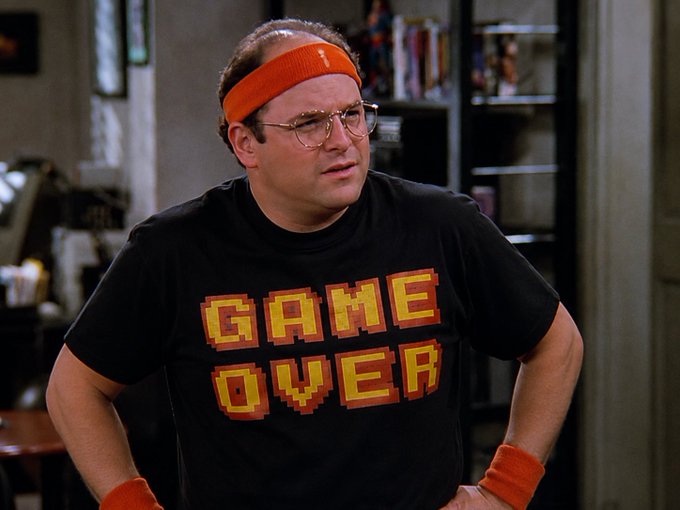 The character George Costanza from Seinfeld wears a black pixel-style t-shirt with "GAME OVER" written in orange and red text, along with an orange headband and wristbands.