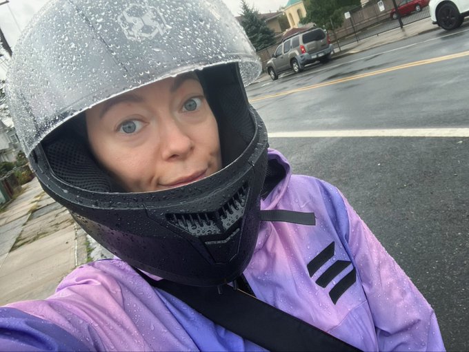 A person wearing a rain-covered silver helmet and a purple Adidas rain jacket takes a selfie on a wet street.