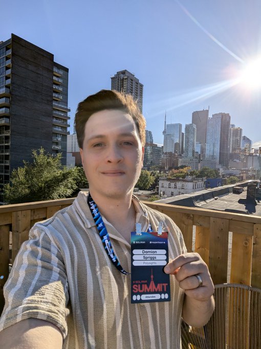A selfie of a young man named "Dominion Springs" holding a "SOLANA SUMMIT" badge, taken on a rooftop with a city skyline and bright sun in the background.