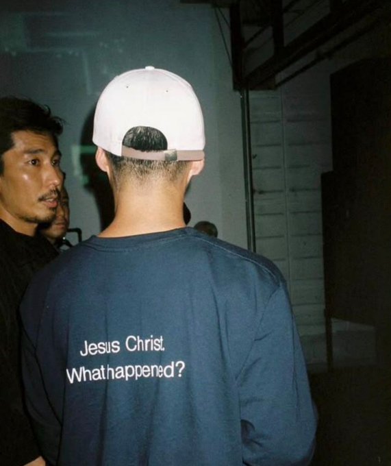 A man seen from behind wearing a white cap and a navy blue long-sleeve shirt printed with 'Jesus Christ What happened?'