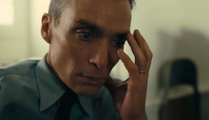 A close-up shot of a man with short hair wearing a light blue shirt and dark tie, holding his hand to his forehead in a distressed or contemplative expression.