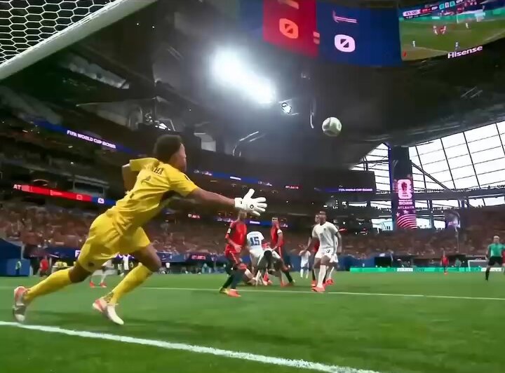The image shows a soccer game taking place with players wearing yellow jerseys engaged in an intense match. One player has just kicked the ball towards the goal, which is located near the top of the image. The goalkeeper is diving to make a save, while other players are scattered across the field. In the background, there's a large screen displaying the score and additional information about the game. A person in the foreground can be seen wearing a yellow shirt with the word "bitcoin" written on it. This individual appears to be involved in some sort of financial transaction related to the soccer match or is simply observing the game from the sidelines.
