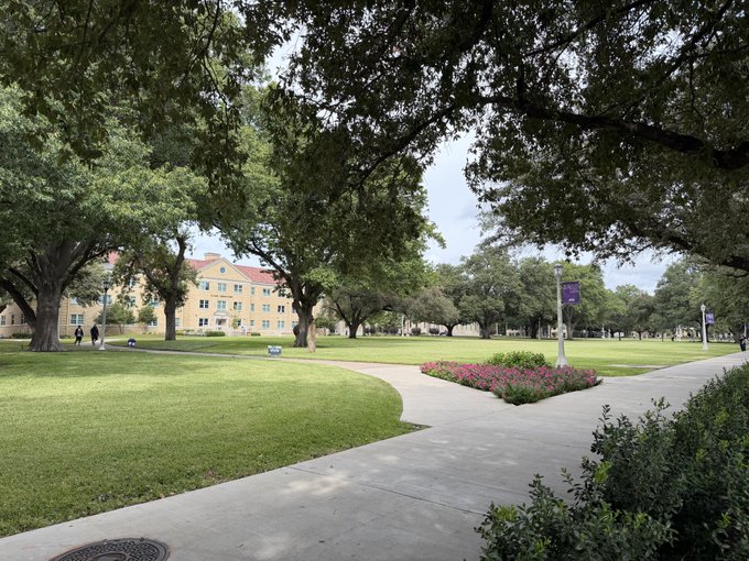 A scenic view of a university or college campus quad with large mature trees, green lawns, concrete walkways, pink flower beds, and a yellow multi-story building in the background under a partly cloudy sky.