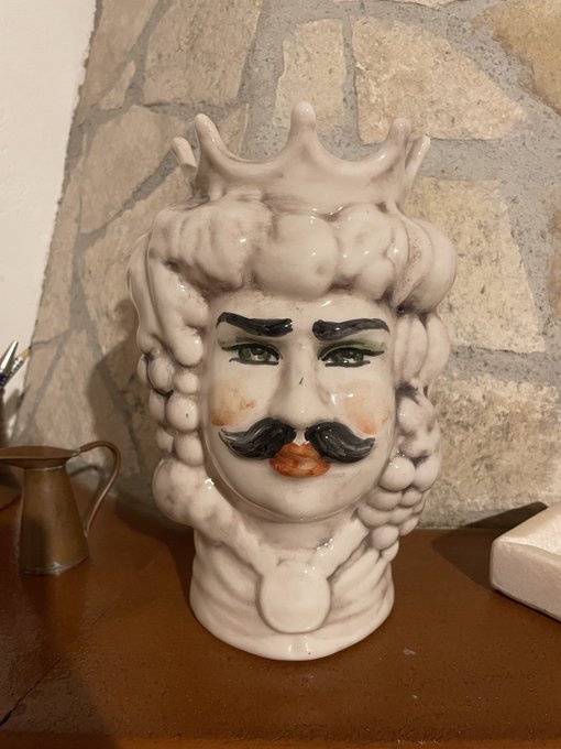 A ceramic decorative pitcher shaped like a king's head with a crown, prominent mustache, and wild hair, displayed against a stone wall.