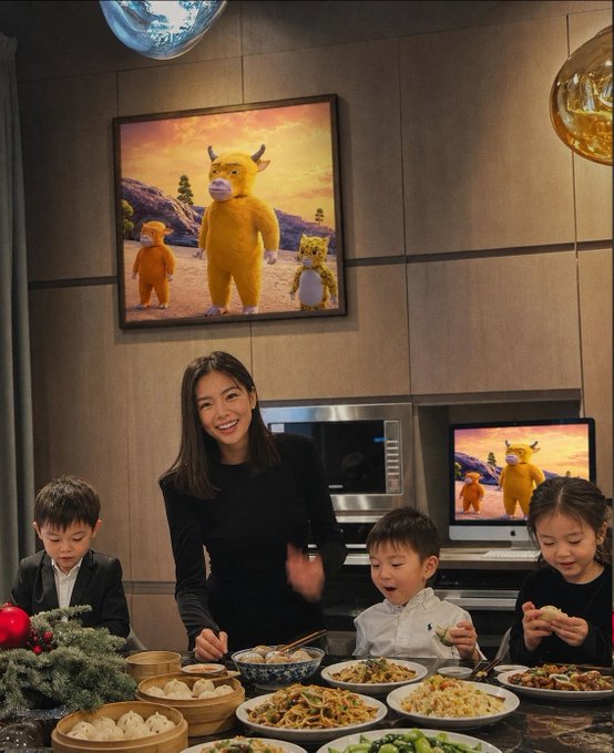 A smiling woman and three children gather around a feast of dumplings and dishes in an elegant kitchen, with a large painting of a yellow cow character and a matching screen on the wall behind them. The whimsical meme ties to the post's punchline about a "Niu Lai" memecoin (niu meaning "cow" in Chinese), humorously suggesting the family's wealth came from investing in this cartoon cow-themed token.