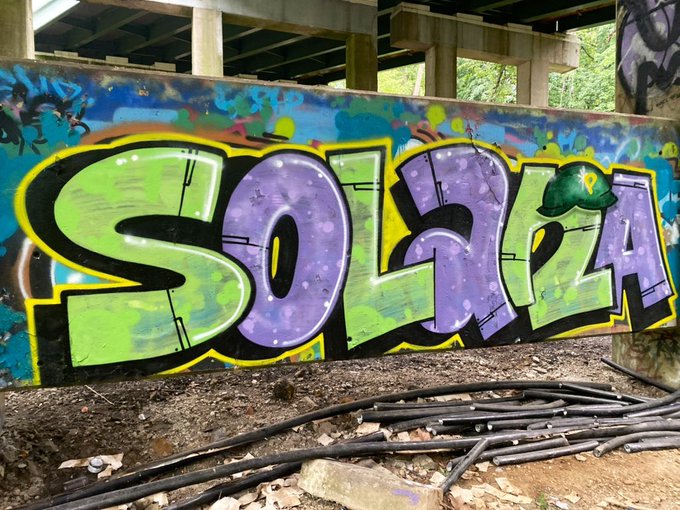 The image features a graffiti-covered wall with the word "Solana" written in purple, green, and yellow letters. The text appears to be spray painted onto the wall, giving it an artistic and urban feel. The surrounding area includes some rocks and pipes, adding to the overall atmosphere of the scene. The post that reads "Is @Solana" is located near this graffiti-covered wall, suggesting a connection between the graffiti art and the name of the person who created or found the image.
