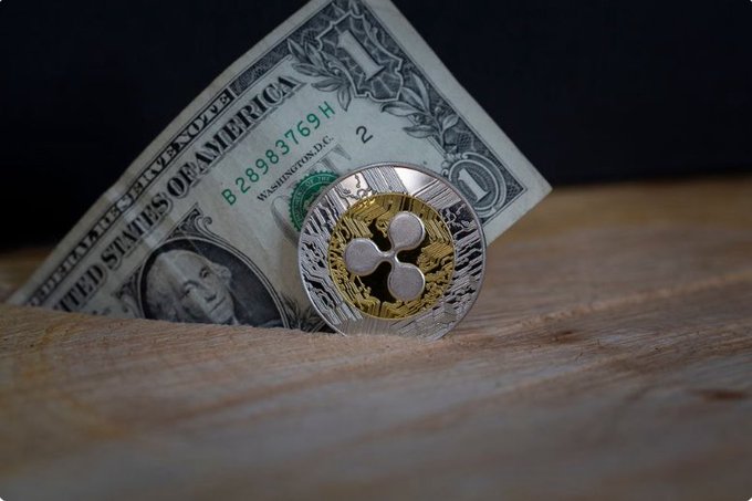 A physical XRP cryptocurrency coin resting against a partially folded United States one-dollar bill on a wooden surface against a dark background.