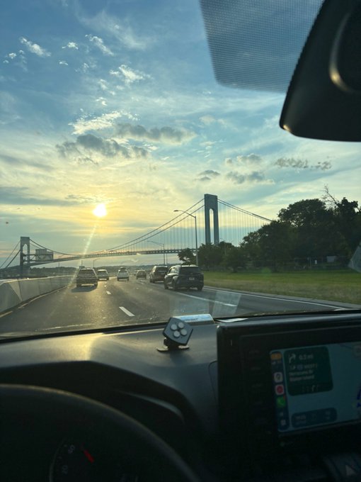 View from inside a car driving on a highway toward a suspension bridge at sunset, with a music player visible on the dashboard.