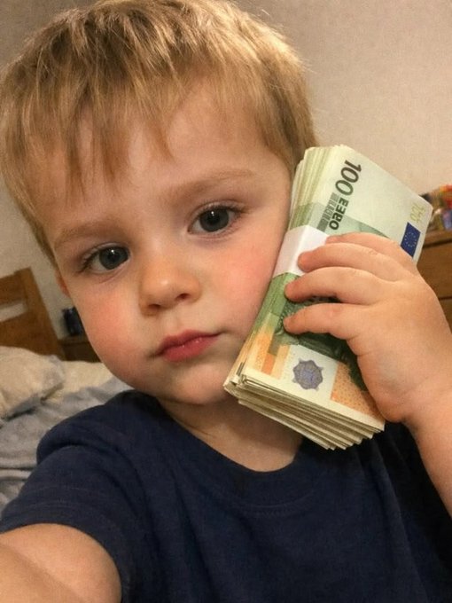 A young blonde child holds a thick stack of 100 Euro banknotes against their cheek, looking at the camera with a serious expression.