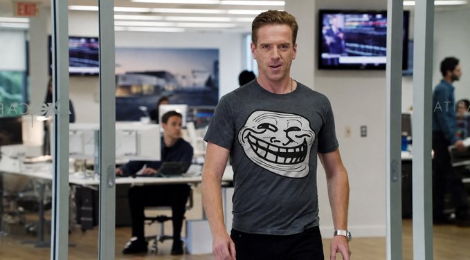 A man wearing a gray t-shirt featuring a troll face meme walks through a modern office environment.