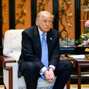 The image features a man wearing a suit, sitting in an armchair with his hands clasped together. He appears to be a political figure or a public official, as suggested by the caption "let' s try bringing back Mr. President". The presence of flowers and potted plants around him further emphasizes the importance of this event.