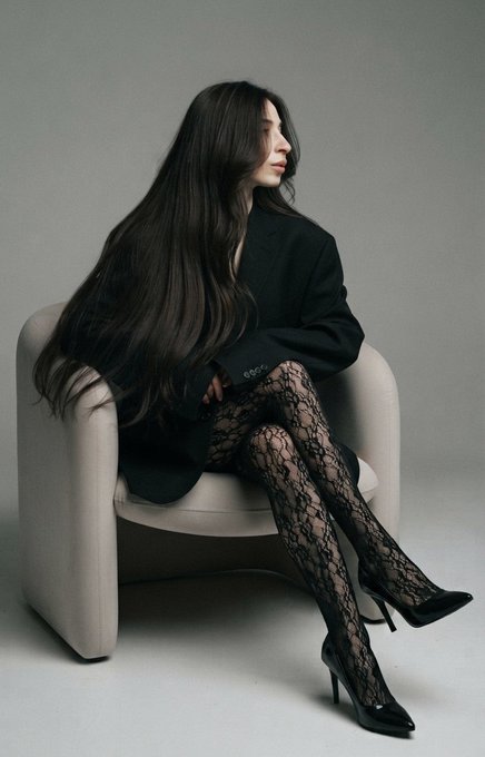 A fashion photo of a woman with very long black hair sitting in a beige armchair, wearing an oversized black blazer and black lace patterned tights with pointed black stiletto heels.
