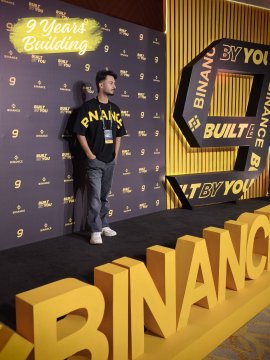 A man wearing a black Binance t-shirt standing in front of a yellow 'BINANCE' step-and-repeat backdrop with '9 Years Building' signage, suggesting a Binance anniversary event.