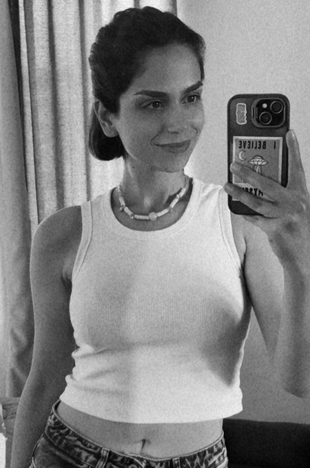 A black-and-white mirror selfie of a smiling woman wearing a white tank top and beaded necklace, holding up a phone with a case displaying 'BELIEVE.'