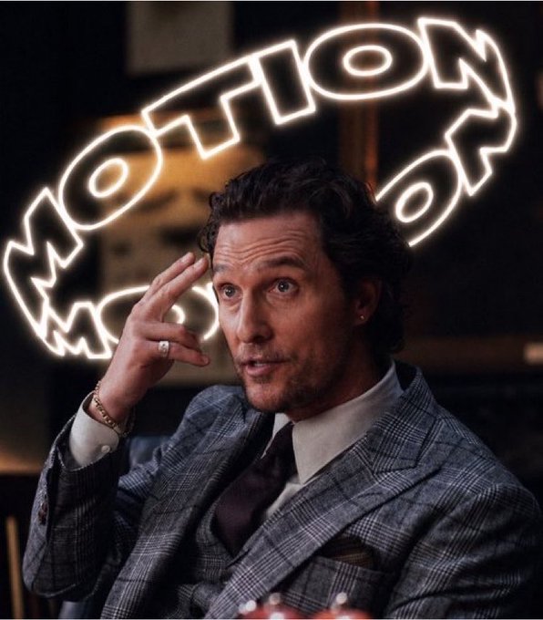 A man in a plaid suit speaks while gesturing near his temple, with a glowing neon 'MOTION' sign behind him.
