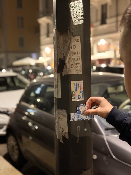 A hand applies a penguin-shaped sticker labeled 'PENGU' to a lamppost covered in old torn stickers, including ones reading 'WIZARD' and 'HACK CLUB', with parked cars and a nighttime street scene in the background.