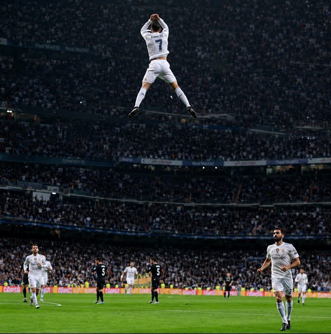 Soccer player Cristiano Ronaldo, wearing a white Real Madrid jersey with 'RONALDO 7' on the back, performs a jumping celebration in a packed stadium.