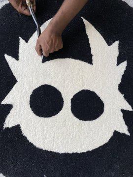 Hands use scissors to trim the edges of a white shaggy rug shaped like a spiky-haired cartoon character's head with two black eye holes.