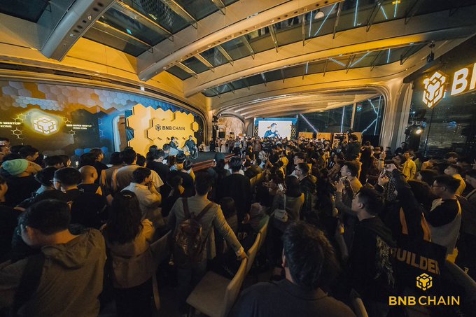 A large crowd gathers at a BNB Chain event, with a stage displaying the BNB Chain logo and branding visible on screens and signs.
