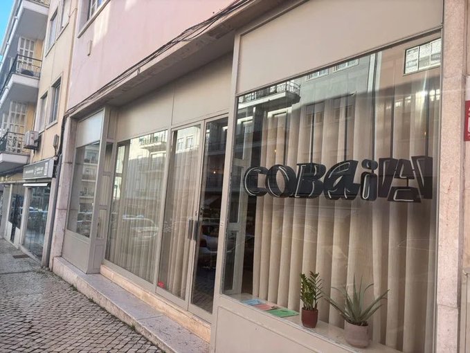 Street view photograph of a storefront with large windows displaying the stylized logo 'COBAÏA' in reverse lettering, with potted plants visible inside and apartment buildings reflected in the glass.