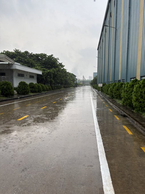A wet, empty road stretching between a small building with a row of bushes on the left and a large blue industrial warehouse on the right under an overcast sky.