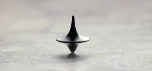 The image features a black object, possibly a toy or a piece of art, placed on a gray surface. The object has a unique design, with a pointed end that resembles an upside-down lid or spout. The background appears to be a light brown color, which contrasts with the dark hue of the object and makes it stand out more prominently in the image.