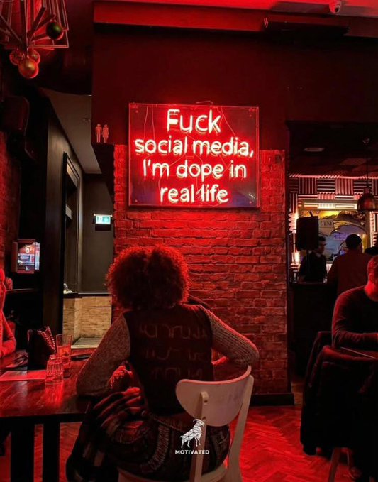 A woman with curly hair sits at a table in a red-lit bar or restaurant beneath a neon sign that reads, 'Fuck social media, I'm dope in real life.'