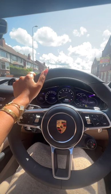 A view from inside a Porsche (identifiable by the steering wheel logo), showing a woman's hand on the wheel with manicured nails, a gold watch, and bracelets, parked on a sunny street.