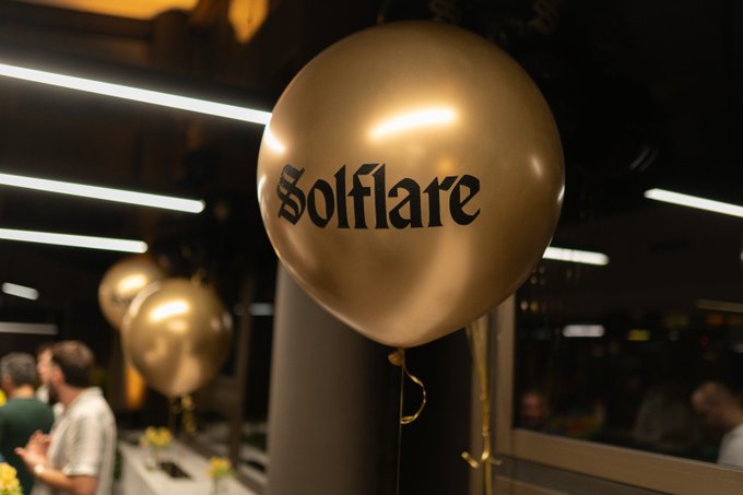 A gold balloon emblazoned with "Solflare" in gothic-style lettering floats at what appears to be a lively corporate event, with blurred figures mingling in the dimly lit background of an office space. The celebratory atmosphere captured in the image reflects the post's description of an unforgettable gathering at Solflare HQ in Belgrade, where connections were made and ideas exchanged among team members.