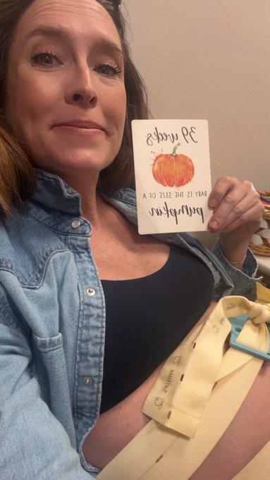 A smiling pregnant woman in a denim shirt holds up a card that reads "thank you, may the size of your blessings be measured by the size of your baby" with a pumpkin illustration, along with a pair of baby socks, celebrating receiving compliments from strangers at 39 weeks of pregnancy.