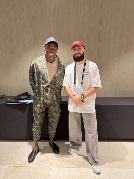 Two men pose together at an event; the man on the left wears a camouflage outfit and grey cap, while the man on the right wears a white t-shirt, red cap, and lanyard.