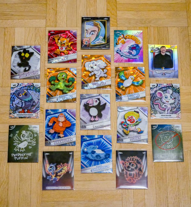 A collection of colorful trading cards or NFT collectibles spread out on a wooden floor, featuring various cartoon characters including Pepe the Frog, a devil, elephant, duck, clown, and other meme characters.