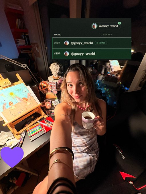 A woman in a striped dress takes a selfie while holding a mug in her art studio; an overlay shows the Instagram handle '@gozyy_world' ranked at #207.