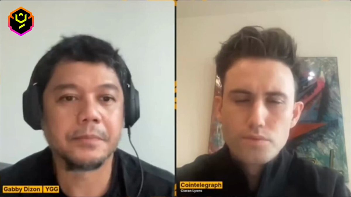 The image shows two men wearing headphones, both looking at their phones or computers. The man on the left has his phone open, while the other man on the right also has his phone open. They are likely engaged in a conversation or browsing content online.