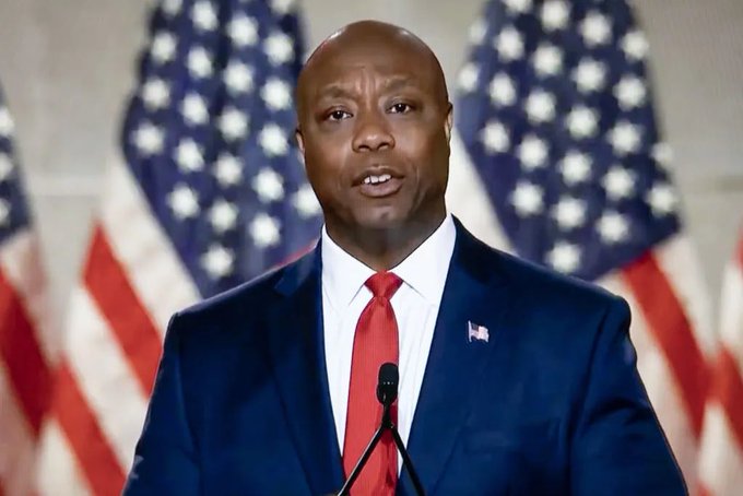 Senator Tim Scott, dressed in a dark suit with a red tie and an American flag lapel pin, speaks at a podium with microphones against a backdrop of multiple American flags, delivering a message urging the Senate to vote on crypto market structure legislation in July.