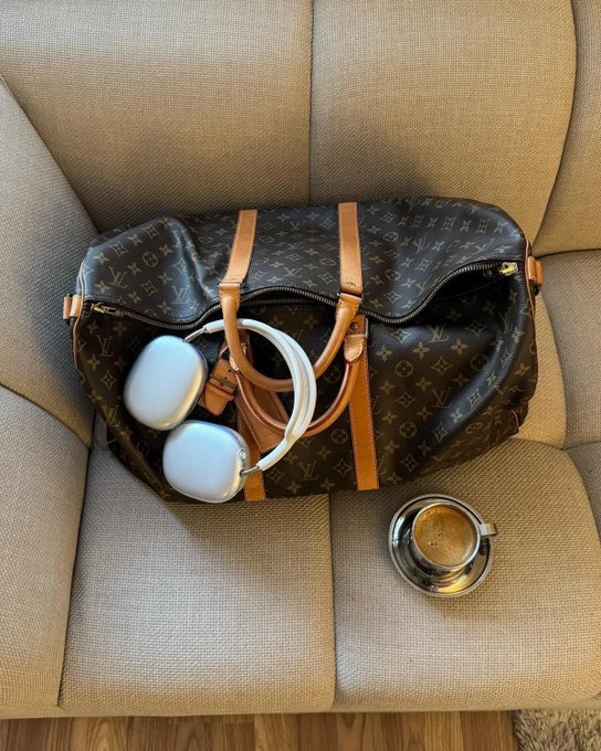 A Louis Vuitton monogram duffle bag sitting on a beige chair, with light blue over-ear headphones resting on top and a metal cup beside it.