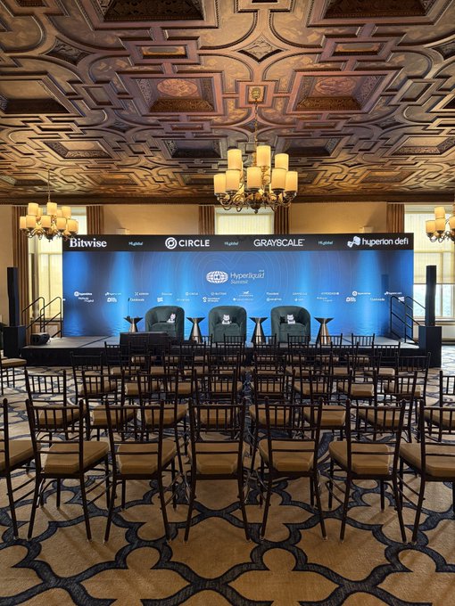 A panel stage set up at the Hyperliquid Summit features branding from sponsors including Bitwise, Circle, Grayscale, and Hyperliquid Delphi, surrounded by rows of empty chairs in an elegant ballroom with ornate ceilings and chandeliers at the New York Athletic Club. The image illustrates the post's theme by capturing the stark contrast between the lavish, fully arranged venue and the empty audience seats—a fitting visual metaphor for a crypto bear market, where even high-profile industry events draw sparse attendance despite heavy investment in production and sponsorship.