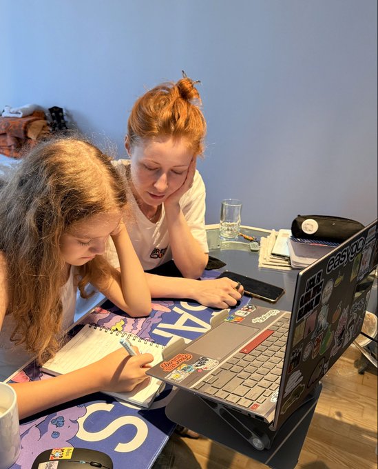 Two people, one with curly reddish-brown hair and another with red hair in a bun, sit at a table working on a laptop covered in stickers while taking notes in a notebook.