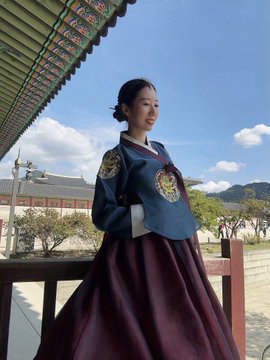 A young woman stands smiling at what appears to be a Korean palace, wearing a traditional hanbok with a navy blue jeogori top featuring dragon embroidery and a maroon chima skirt.