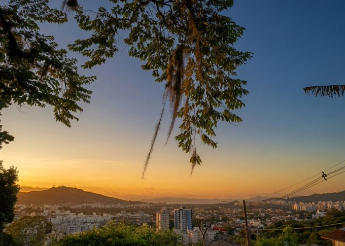 A scenic sunset photograph captures a cityscape nestled in a valley surrounded by hills, framed by tree branches with hanging moss in the foreground under a gradient orange and blue sky.