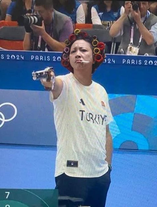 A female Turkish athlete at the Paris 2024 Olympics wearing a 'TÜRKİYE' jersey and hair rollers, pointing a pistol at the camera while smoking a cigarette.