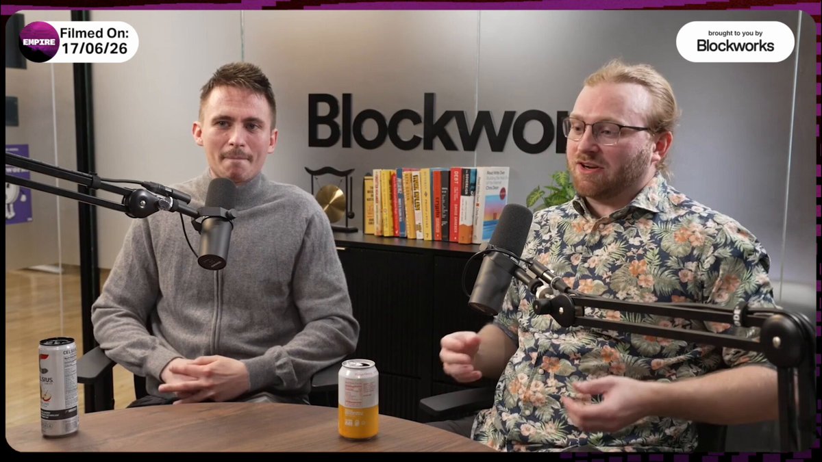 In this image, there are two men sitting at a table with microphones in front of them. They appear to be having a conversation or participating in an interview. The background features a bookshelf filled with numerous books, suggesting that they might be discussing topics related to finance and investing. One man is wearing glasses while the other has a beard.