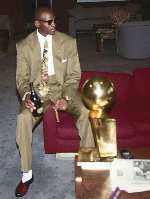 A man in a tan suit and sunglasses sitting on a red sofa holding a champagne bottle, with a golden trophy resembling the Larry O'Brien NBA Championship Trophy beside him.